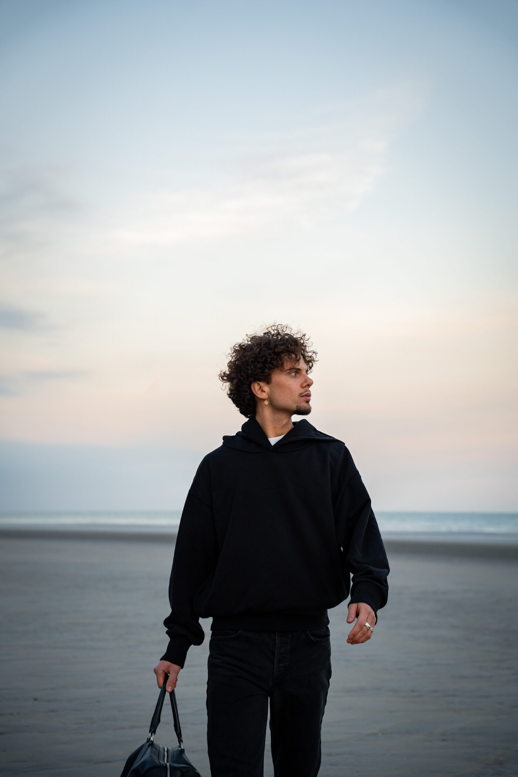 Young man with curly hair in a black hoodie walks on an empty beach at dusk, carrying a bag. The sky is soft with pastel hues, evoking a calm, contemplative mood.