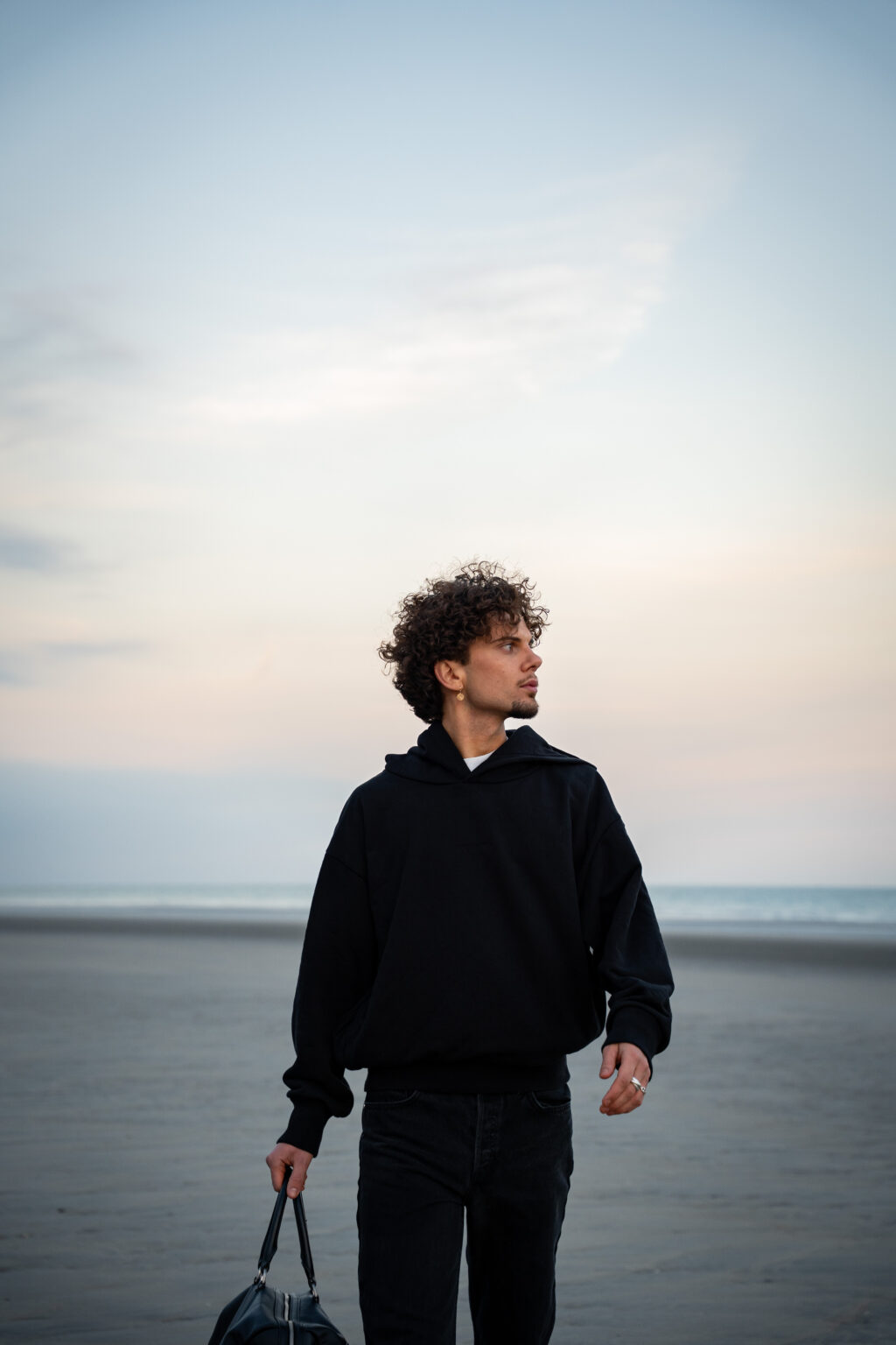 Young man with curly hair in a black hoodie walks on an empty beach at dusk, carrying a bag. The sky is soft with pastel hues, evoking a calm, contemplative mood.