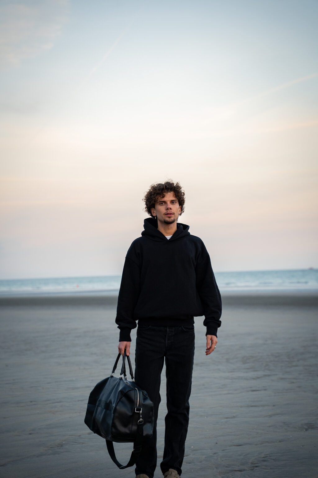 Young man with curly hair in a black hoodie stands on a beach at sunrise, holding a large travel bag. He looks contemplative against the calm ocean backdrop.