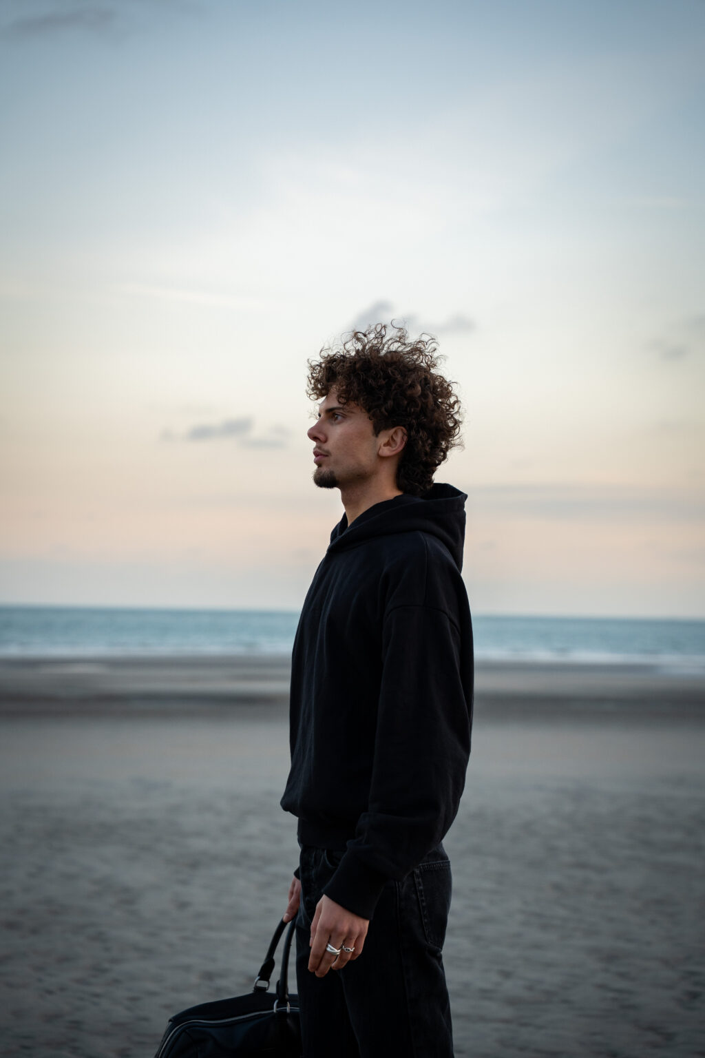 A young man with curly hair stands on a beach at dusk, wearing a black hoodie and holding a bag. He gazes toward the horizon, conveying a contemplative mood.