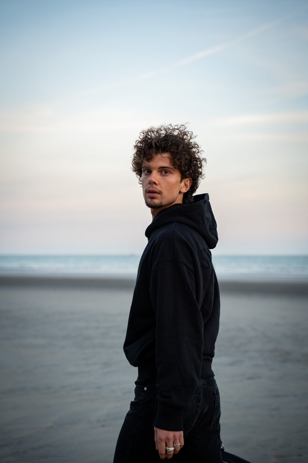 Young man with curly hair wearing a black hoodie looks back on a deserted beach at dusk. Soft pastel sky and calm ocean create a contemplative mood.