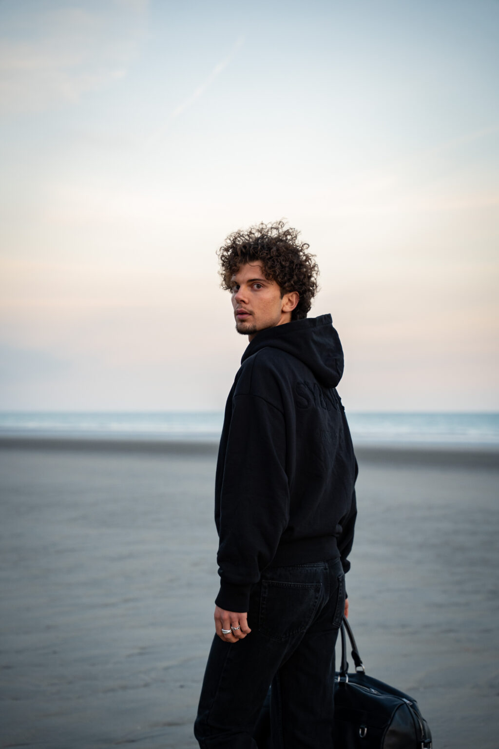 Young man with curly hair in a black hoodie stands on a serene beach at dusk, holding a bag. He gazes thoughtfully over his shoulder.