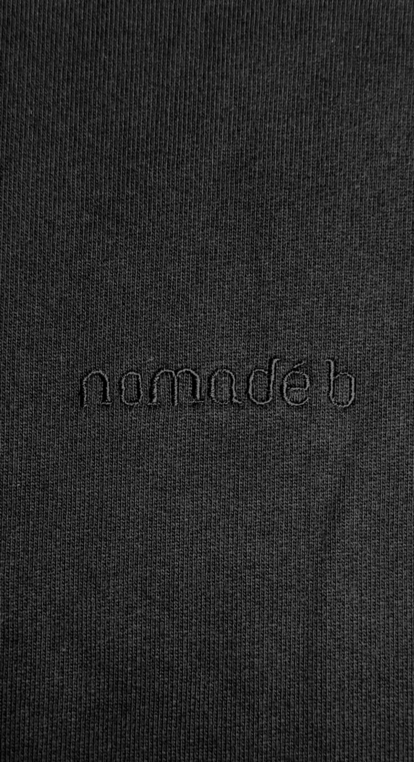 Close-up of a black fabric with the word "nomadé b" subtly embroidered in matching threads. The tone is minimalist and sleek, emphasizing texture.
